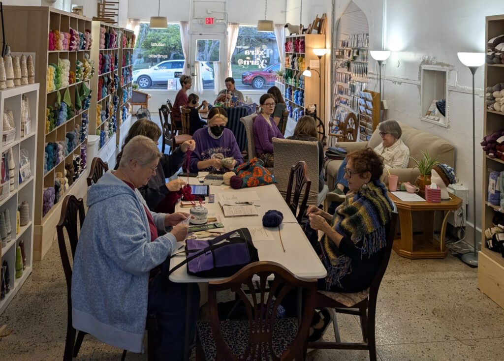 A group of 11 people seated around a two tables on on a sofa all working on fiber arts projects like knitting or crocheting.