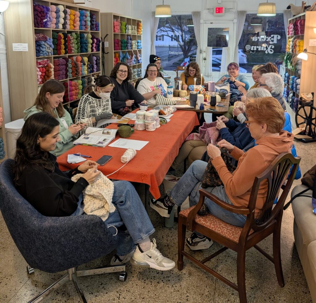 A group of 13 people seated around a table all working on fiber arts projects like knitting or crocheting.