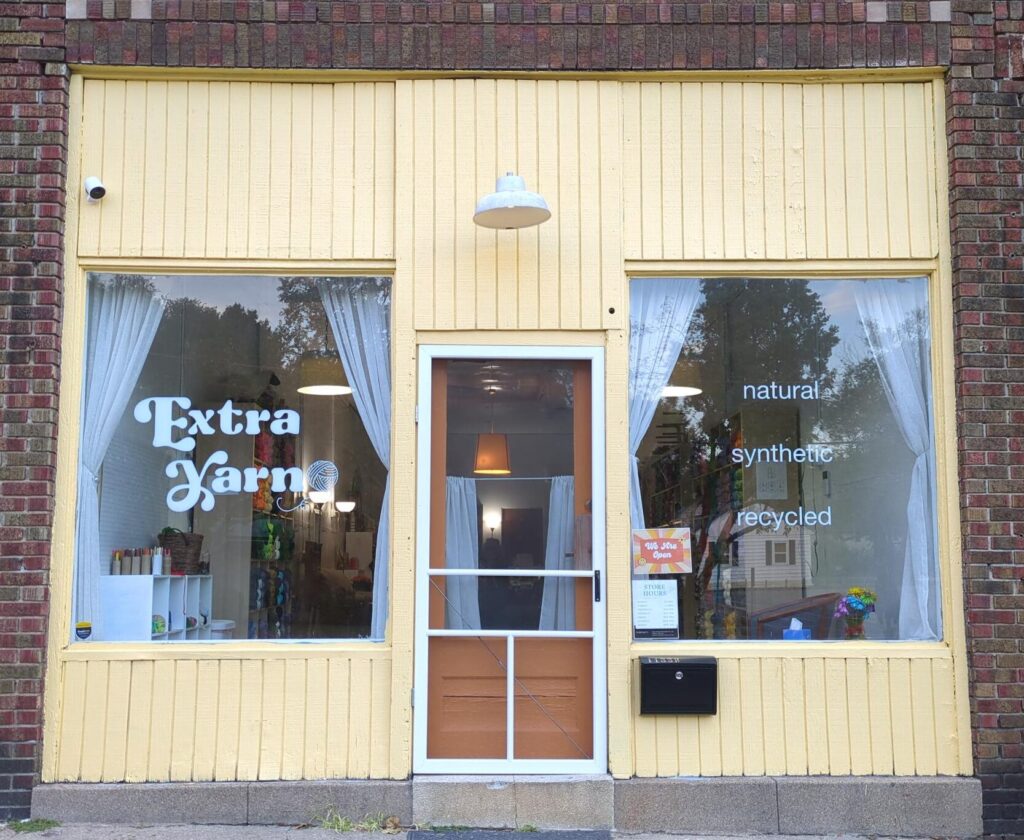 The exterior of Extra Yarn is pastel yellow with an orange front door nestled between two large picture windows.