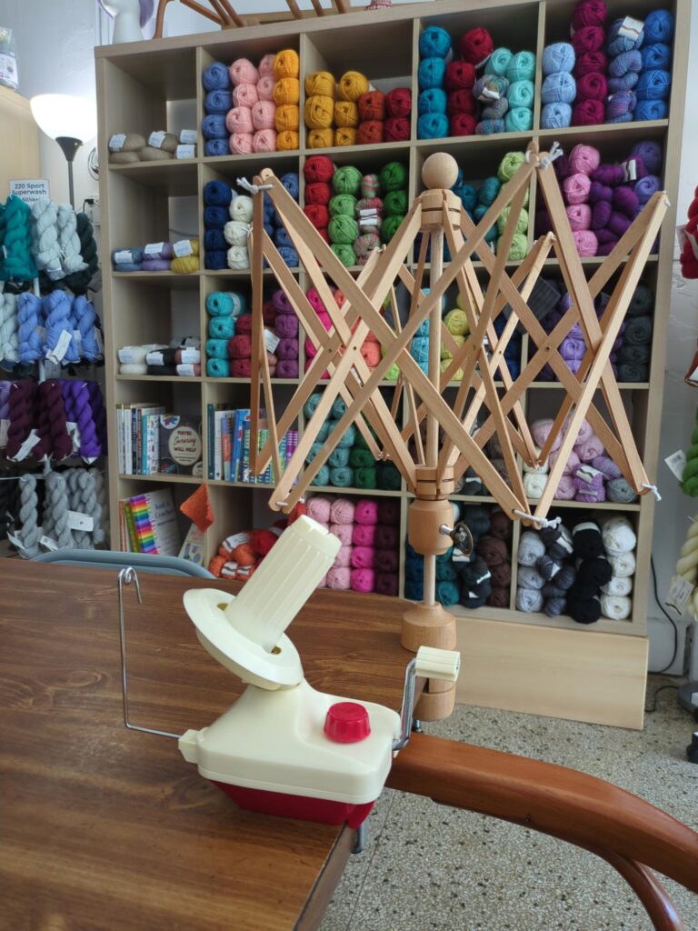 Wooden yarn swift and ball winder attached to a table with a shelf full of colorful yarn in the background.
